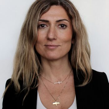 Aralia Eriksson, long brown hair, olive skin wearing black suit and white shirt with two gold chains