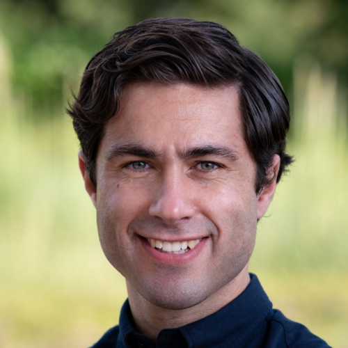 Oli Goodarzi smiling at camera wearing blue shirt and with greenery in the background.