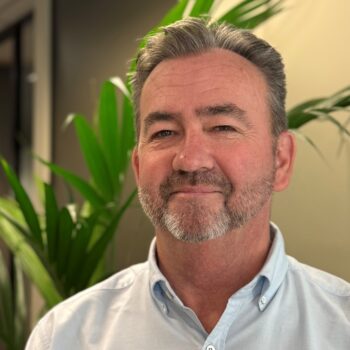 Mark Burns in pale blue shirt stands in the office in front of an indoor plant.
